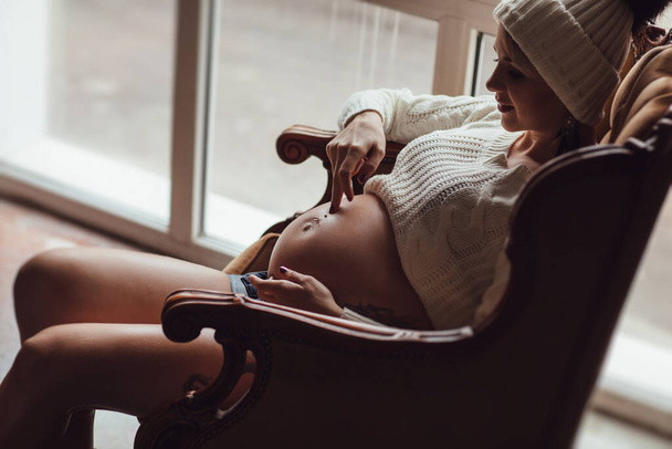 pregnant girl with tattoo in knitted cap sitting in armchair in front of window. - Foto, Imagen