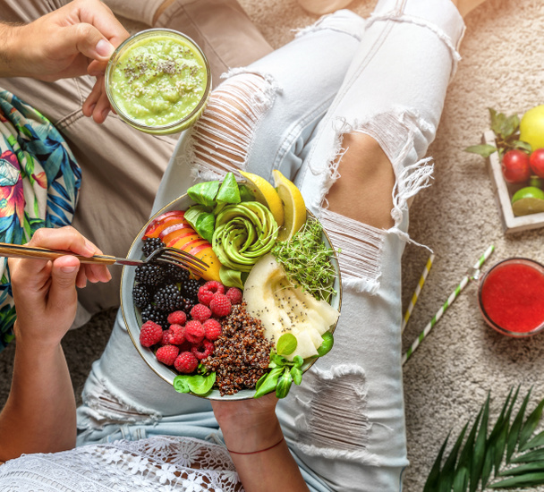 Пара в літньому одязі тримає свіжий салат з кіокою, персиком, зеленню, авокадо, ягодами, динею в мисці на світлому фоні з тропічним листям. Здорова їжа, чисте харчування, миска Будди, вид зверху
 - Фото, зображення