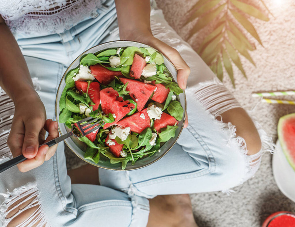 Nő farmerban eszik friss nyári görögdinnye saláta feta sajt smoothie világos háttér trópusi levelek. Egészséges ételek, tiszta evés, Buddha tál saláta, felülnézet, tonizálás - Fotó, kép