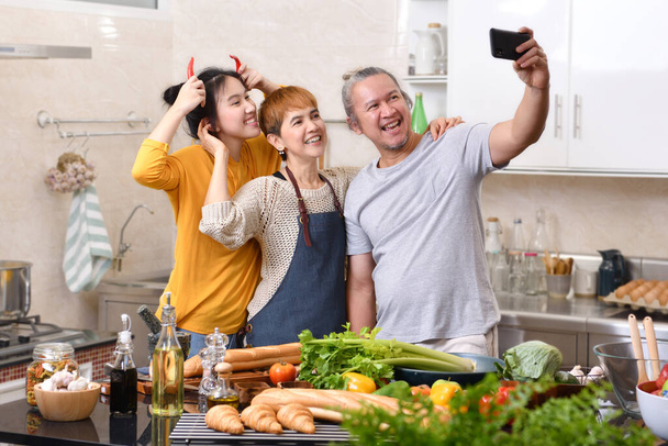 Familia feliz de madre padre e hija cocinando en la cocina haciendo comida saludable juntos y usando un teléfono inteligente para tomar selfies
 - Foto, Imagen