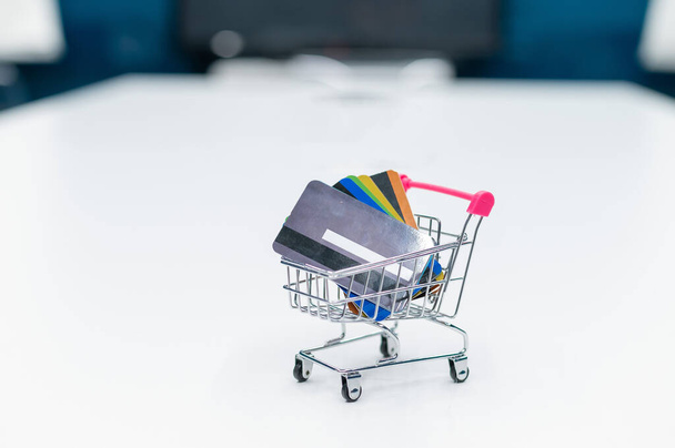 A mini shopping trolley full of credit cards stands on a white table. No people. Close-up of a cart with several different bank debit cards. - Foto, Imagem