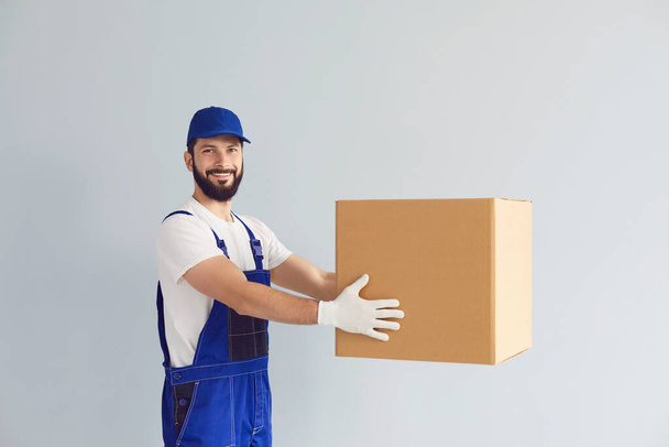 Happy young deliveryman holding cardboard parcel box with mockup for your design against grey background - Photo, image
