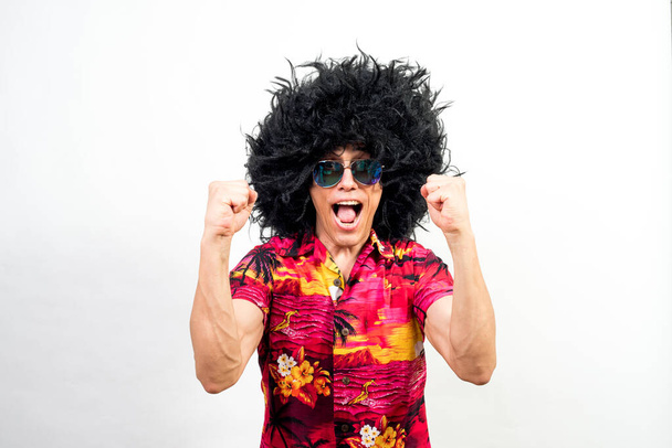 Homem com peruca afro, óculos de sol e camisa havaiana celebrando algo, muito feliz. A meio do tiro. Fundo branco
. - Foto, Imagem