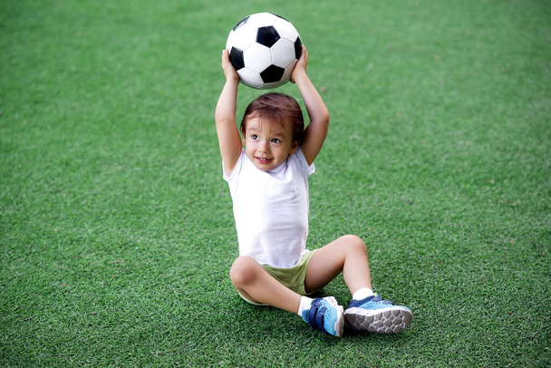 Toddler boy in sports uniform sitting on green footall field holding soccer ball above head. Little child playing with ball on grass. Active childhood and outdoor games for children concept - Foto, imagen