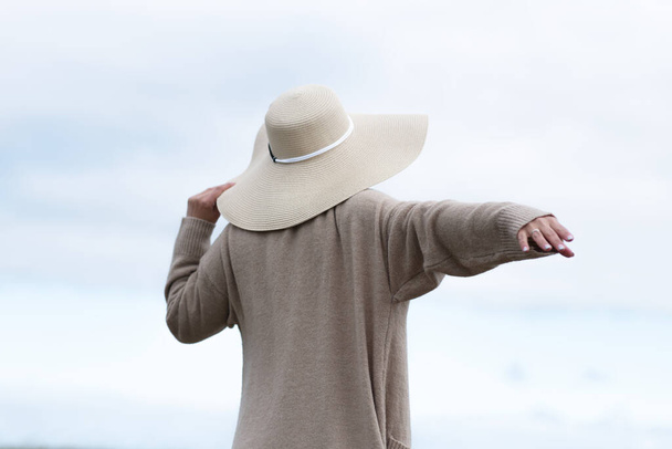 A woman in a hat and a long beige cardigan stands with outstretched arms against the skyhed arms against the sky. High quality photo - Fotó, kép