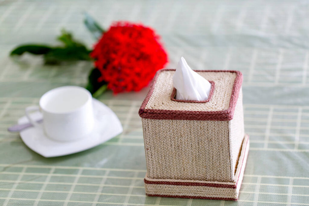 Raaka kuidun väri käsintehty lautasliina laatikko ruokapöydälle, jossa on punainen kukka ja valkoinen teekuppi. Kudospaperilaatikko ravintolassa. - Valokuva, kuva