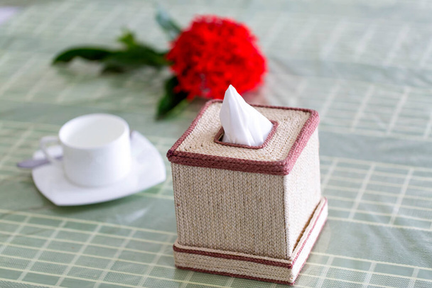 Nyers rost színű kézzel készített szalvéta doboz az ebédlőasztalon egy piros virág és egy fehér teáscsésze. Papírdoboz az étteremről. - Fotó, kép