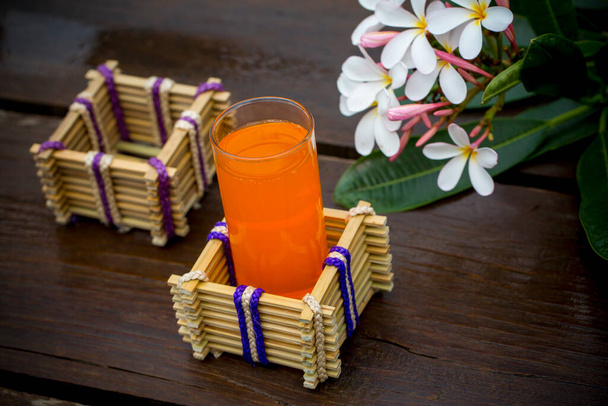 Ένα ποτήρι χυμό πορτοκαλιού με βάση από μπαμπού και φυτικές ίνες. Διακοσμητικό ξύλινο τραπέζι με όμορφα λουλούδια Plumeria. Δοχείο γυάλινο κάτοχος. - Φωτογραφία, εικόνα
