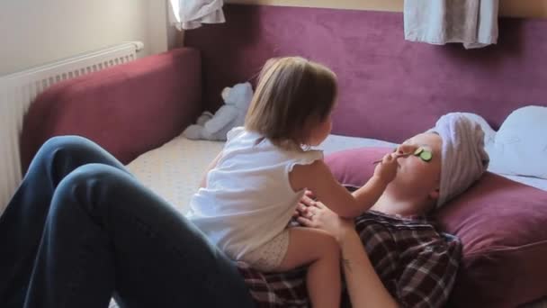 Woman after bath relaxing with cucumbers on eyes. Her little daughter paints Mothers face with orange color. Concept: beauty, family, fun, relax. - Video