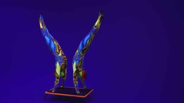 Akrobatická dvojčata v neonovém kostýmu předvádějí stojan na rukou a na hlavě, na modrém cyklorámu v ultrafialovém make-upu v cizím stylu.Světlé fluorescenční těleso září ve tmě. - Záběry, video