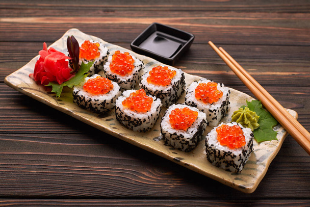 Rollo de sushi con caviar rojo en un plato con wasabi, jengibre, hojas de arce y palillos para sushi, sobre un fondo de madera
 - Foto, Imagen