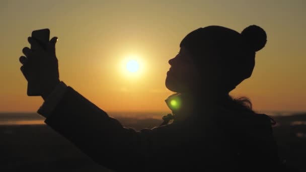 silhouette di una donna al sole, selfie, scattare foto al tramonto, alba. Free Young girl turista blogger registra un selfie video in cima alle montagne utilizzando uno smartphone con un bellissimo paesaggio. - Filmati, video