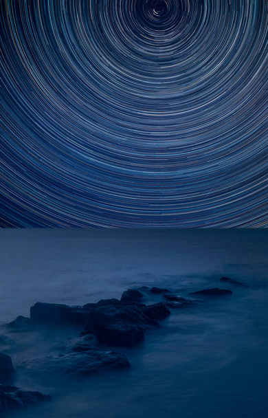 Image composite numérique de sentiers étoilés autour de Polaris avec image de paysage traversant paisible traitée de la mer calme sur les rochers  - Photo, image