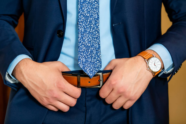 Groom in blue shirt and jacket holding brown belt. Classy blue suit for man. Brown watches on the hand - Foto, afbeelding