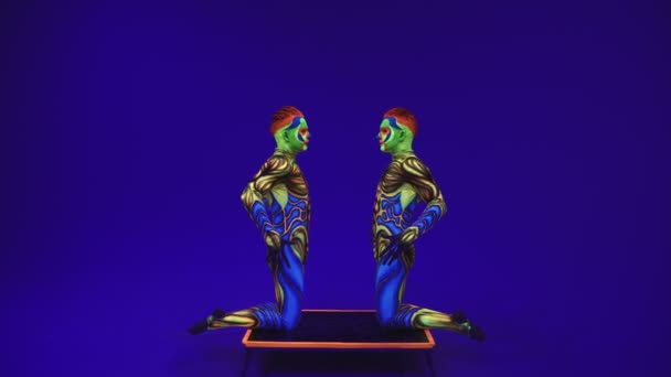 ネオンコスチュメストのアクロバティックな双子は、手と頭の上でスタンドを実行します,エイリアンのスタイルで紫外線メイクで青いサイクロラマに.. - 映像、動画
