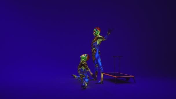 Acrobatische tweeling in neon kostuums om een stand op de handen en het hoofd uit te voeren, op de blauwe cyclorama in ultraviolette make-up in een buitenaardse stijl.Heldere fluorescerende body art gloeit in duisternis. - Video