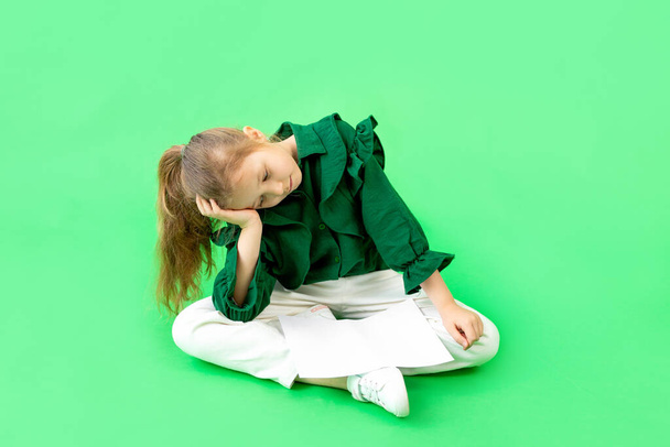 ένα κορίτσι 8 ετών κάθεται σε ένα πράσινο φόντο και κρατά το κεφάλι της - Φωτογραφία, εικόνα