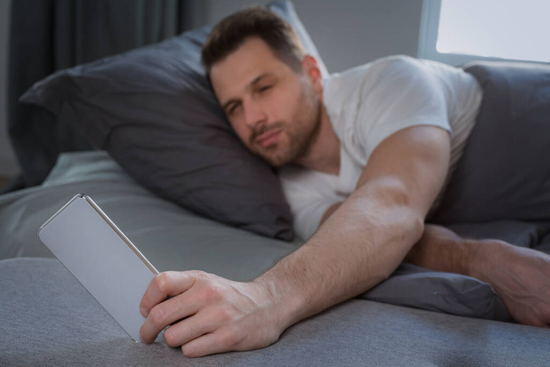 Ο άνθρωπος που χρησιμοποιεί Smartphone αφύπνισης ξαπλωμένος στο κρεβάτι στο σπίτι - Φωτογραφία, εικόνα