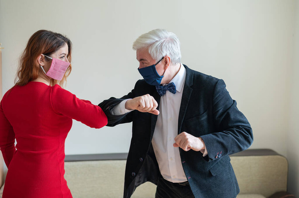 A mature gray-haired man in a suit and a red-haired woman in a red dress wearing in medical masks maintain a social distance and greet their elbows. An alternative to shaking hands and hugs. - Φωτογραφία, εικόνα