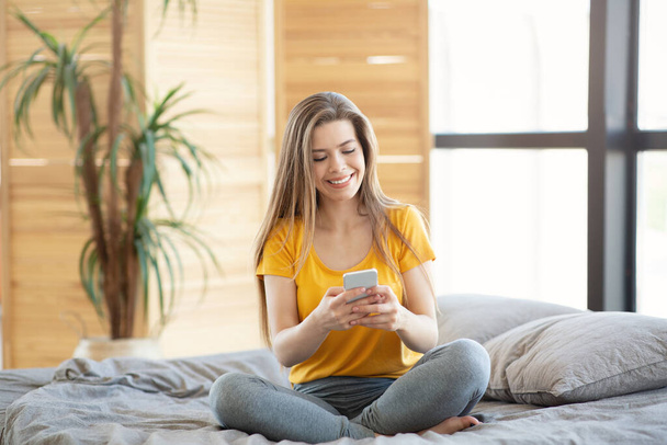 Pretty lady using mobile phone for online communication, work or entertainment on bed indoors - Photo, image