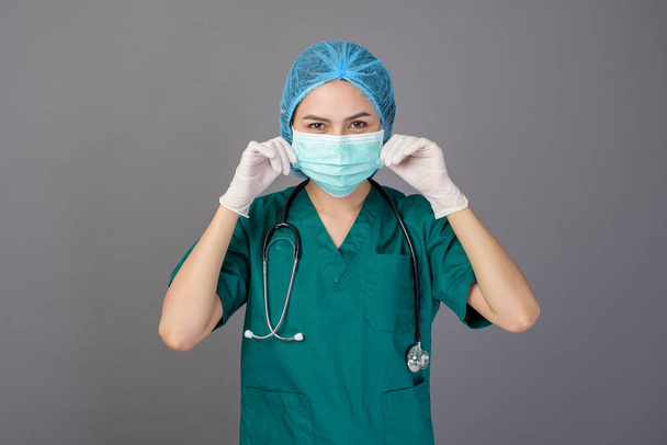a young confident woman doctor in green scrubs is wearing surgical mask over grey background studio - Fotoğraf, Görsel