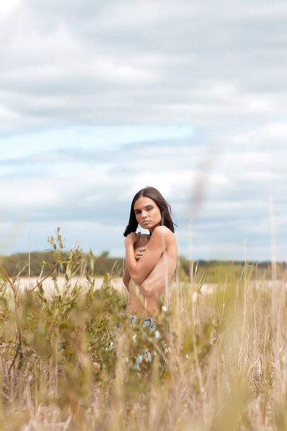Boční pohled nahé brunetky pokrývající prsa a dívající se na kameru přes rameno, zatímco stojí v křoví v přírodě - Fotografie, Obrázek
