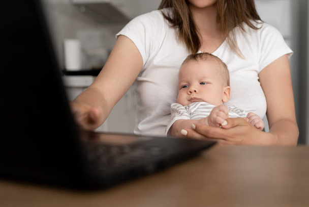 授乳中の赤ちゃんの肖像画を閉じます.その少年は母親の腕の中に座っている。女性はノートパソコンを持って家で働いている。リモート従業員の概念. - 写真・画像