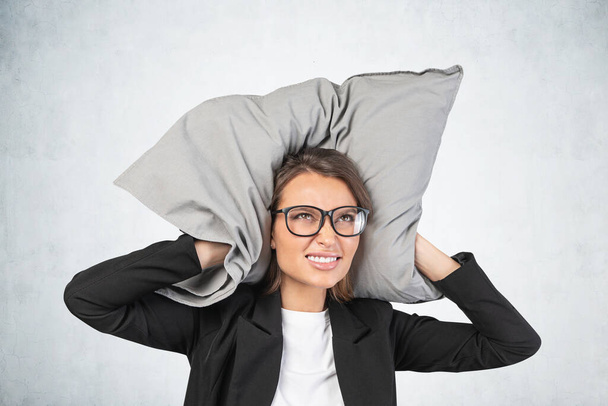 Portrait of frustrated young businesswoman in glasses covering her ears with pillow near concrete wall. Concept of noisy environment and importance of rest - Foto, Imagem