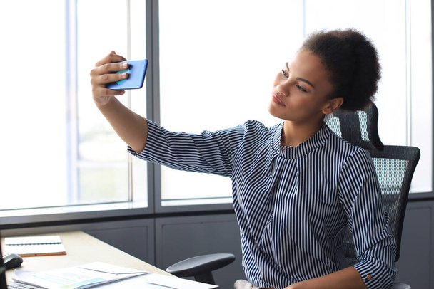 Young african american woman doing selfie for social nets while sitting in creative office - Photo, Image