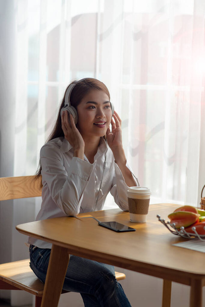 Portrét mladé asijské ženy se baví poslouchat hudbu se sluchátky a horký šálek kávy na stole, šťastný a relaxační čas - Fotografie, Obrázek