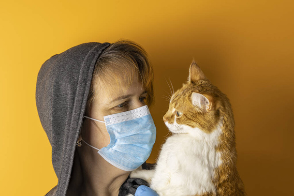 Female in a medical mask and hood looking at red white cat on a yellow background. Health protection during flu virus outbreak, coronavirus epidemic infectious diseases and allergy to animal fur - Foto, imagen