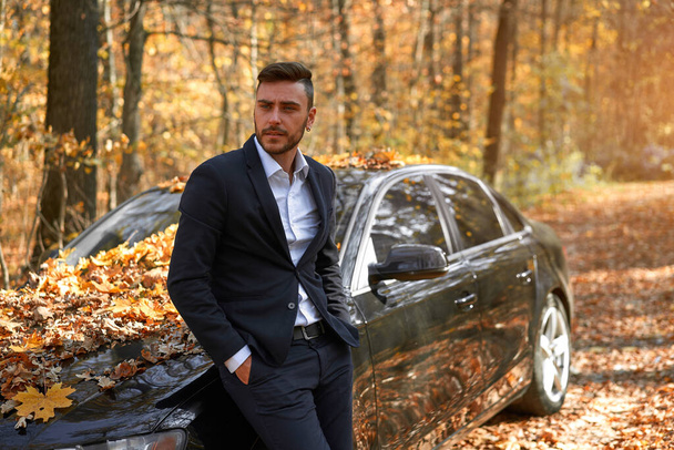A handsome Caucasian man wearing business suit standing near black car on nature.  Young adult businessman travels by car on a beautiful sunny autumn day. Elite luxury driver. - Fotoğraf, Görsel