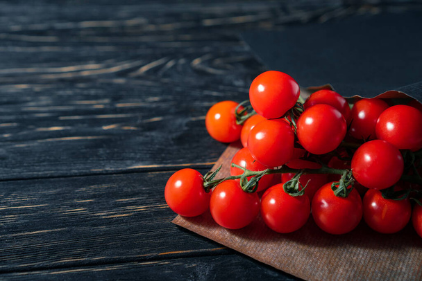 Koyu ahşap masadaki kağıt ambalajda kiraz domatesleri. Siyah arka planda sulu olgun sebzeler. Sağlıklı beslenme. vejetaryen yemeği - Fotoğraf, Görsel