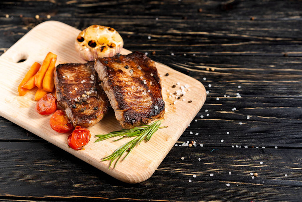 A juicy piece of fried meat with grilled cherry tomatoes garlic and carrot lies on a cutting board against a black wooden table. Degree of roasting well done. Food concept with copy space - Foto, Bild