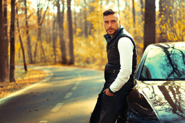 A handsome Caucasian man wearing style black vest standing near black car on nature.  Young adult businessman travels by car on a beautiful sunny autumn day. Elite luxury driver. - Fotoğraf, Görsel