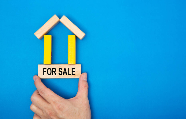 Male hand placing a Buy/Sell sign in a house made of wooden blocks in a conceptual image of real estate market. Over blue background. - Photo, Image