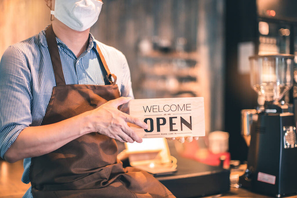 Business owner attractive young Asian man in apron hanging we're open sign on front door  welcoming clients to new cafe. Happy waiter with protective face mask holding open sign while standing at cafe . - Фото, зображення