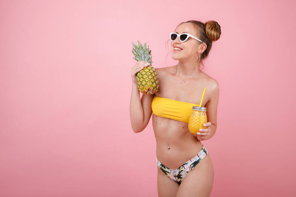 girl in a swimsuit with pineapple and juice on a pink background. summer mood - Photo, Image