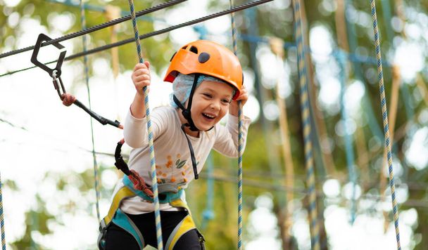 Little girl in helmet climbs ropes in adventure park outdoors. Extreme sport, active leisure on nature. - Fotografie, Obrázek