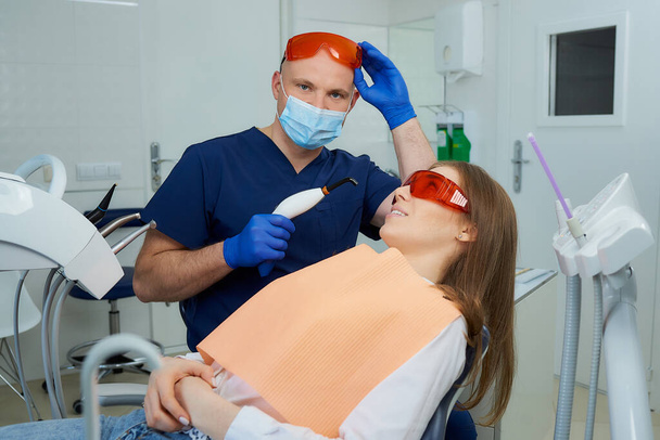 Un dentista con una mascarilla médica y gafas de protección UV sosteniendo una luz de polimerización dental cerca de una chica en la silla dental. Un médico y una paciente antes del tratamiento en el consultorio de un dentista
. - Foto, Imagen