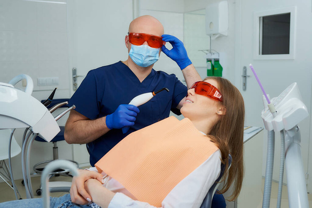 Dentysta w masce medycznej sprawdza okulary ochronne UV i trzyma światło polimeryzacji dentystycznej w pobliżu dziewczyny na krześle dentystycznym. Lekarz i pacjentka podczas leczenia w gabinecie stomatologicznym - Zdjęcie, obraz