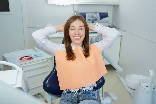 Uma jovem feliz relaxou na cadeira dentária, jogando as mãos atrás da cabeça. Uma paciente do sexo feminino está demonstrando seu excelente sorriso após o tratamento no consultório de um dentista
. - Foto, Imagem
