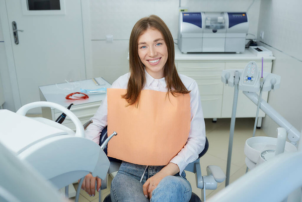Μια χαρούμενη νεαρή γυναίκα χαλαρή στην οδοντιατρική καρέκλα. Μια γυναίκα ασθενής επιδεικνύει το υπέροχο χαμόγελό της μετά τη θεραπεία στο γραφείο ενός οδοντιάτρου.. - Φωτογραφία, εικόνα