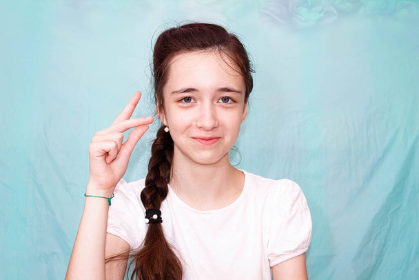 Ein Mädchen mit langem Zopf zeigt mit der Hand den Buchstaben K in der Zeichensprache von Taubstummen. Mittlerer Plan, türkisfarbener Hintergrund. - Foto, Bild