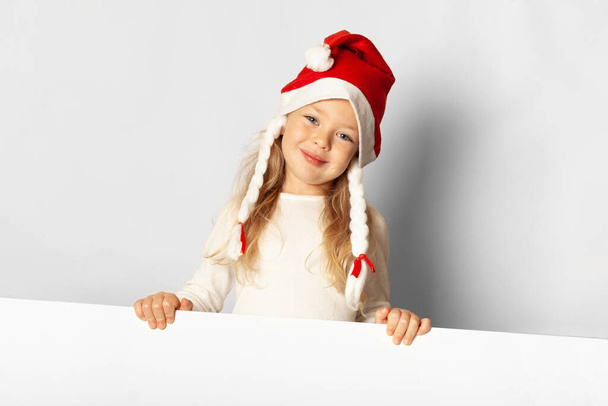 Portrait von kleinen hübschen Mädchen lacht in Weihnachtsmütze mit Verkauf (Werbung) leer. Mit Weihnachtskarton auf weißem Hintergrund. Frohes neues Jahr. - Foto, Bild