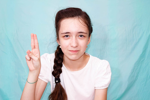 Une fille avec une longue tresse montre la lettre U avec sa main en utilisant le langage des signes des sourds-muets. Plan moyen, fond turquoise. - Photo, image