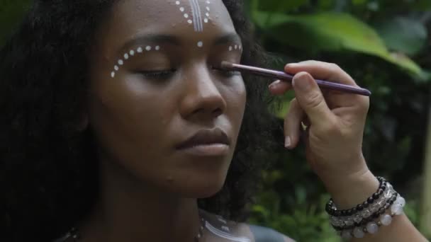 Video of a beautiful young brunette African girl with makeup and painted ethnic lines on the body sitting in a tropical garden - Filmati, video