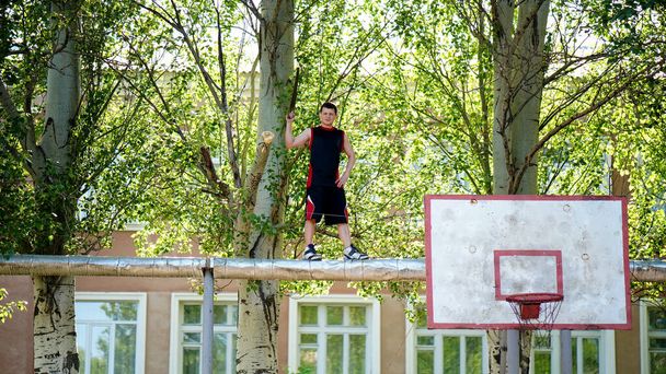 Young man trimming poplar branches over a summer basketball court. Guy standing on pipes high above the ground with a hacksaw in his hand - Photo, Image