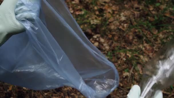 Egy kesztyűs fiatalember kezei egy műanyag üveget tesznek a szemeteszsákba. Szemétgyűjtés az erdőben. - Felvétel, videó