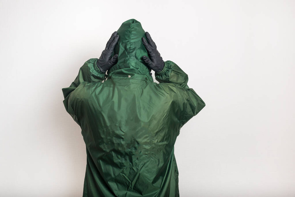 A man in a protective green jacket and black latex gloves stands back to the camera on a white background. Holds the hood with his hands - Φωτογραφία, εικόνα